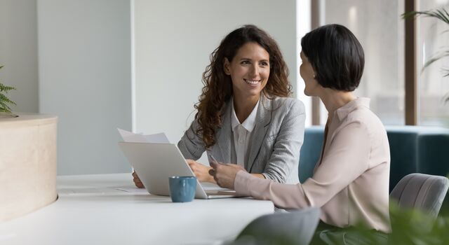 COLOUR-IMAGE_two-women-smiling-whilst-at-an-office-desk_jul26